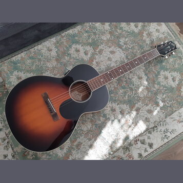 Akustická kytara s vyřezem v těle a tmavým, přechodovým lakem (sunburst) od černé k hnědé. Na hlavě je viditelné logo "Epiphone". Uvnitř otvoru kytar…