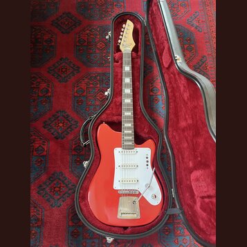 Červená elektrická kytara v černém tvrdém pouzdře s červeným vnitřkem. Nástroj má bílý pickguard, tři snímače a chromovaný most s tremolem. Na hlavě…
