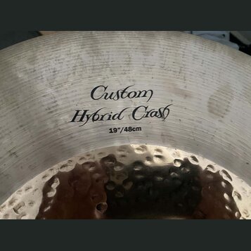Detailní záběr na povrch činchy z kovu. Na povrchu je černý nápis „Custom Hybrid Crash“ a pod ním označení „19"/48cm“. Levá část fotky ukazuje kovaný…