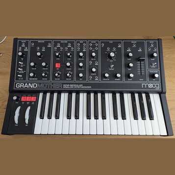 Černý semi-modulární analogový syntetizátor s bílými klávesami. Na předním panelu je nápis GRANDMOTHER SEMI-MODULAR ANALOG SYNTHESIZER a logo moog. P…
