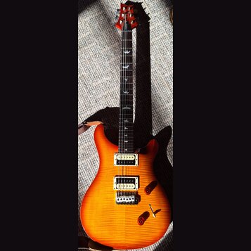 Elektrická kytara s žlutě-oranžovým přechodem (sunburst) a viditnou strukturou dřeva. Na hlavě je červený lak a bílý nápis PRS. Hmatník má vložky ve…