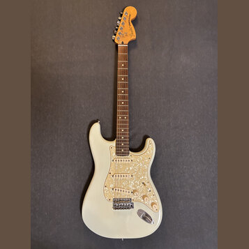 Elektrická kytara s bílým trupem a hmatníkem z tmavého dřeva. Na hlavě je nápis Fender STRATOCASTER. Kytara má tři snímače a plech s perleťovým vzore…
