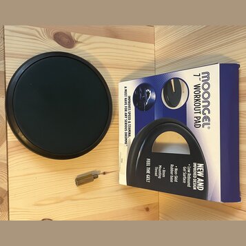 Černý tréninkový pad na bubny s kulatým tvarem. Vedle něj je modrá krabice s nápisy MOONGEL®, 7" WORKOUT PAD, FEEL THE GEL!, IMPROVES SPEED & STAMINA…