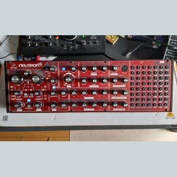 Červený semi-modulární syntetizátor s nápisy behringer a NEUTRON. Přední panel obsahuje zahlreichetočítka, přepínače a patchovací pole s označením IN…