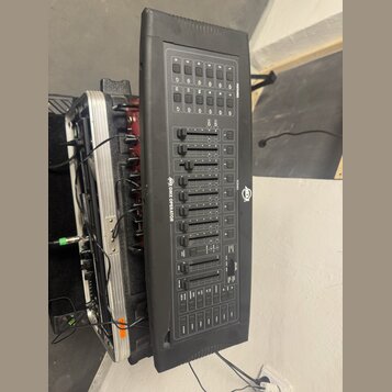 Černý DMX kontrolér s ovládací plochou obsahující posuvníky, tlačítka a displeje. Na přístroji jsou viditelné nápisy DMX OPERATOR a SCENE1, doprováze…