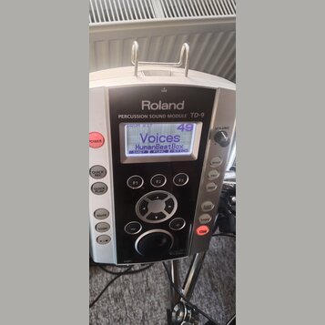 Stříbrný perkusní zvukový modul značky Roland, model TD-9. Na displeji je zobrazen text DRUM KIT 49, Voices a HumanBeatBox s dalšími nápisy INST, FUN…
