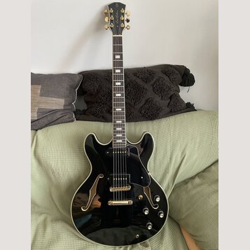 Černá polodublová elektrická kytara s lesklým povrchem a f-otvory. Na hlavě hmatníku je zlatý nápis "Joe Pass". Nástroj má zlaté ladičky, černý pickg…