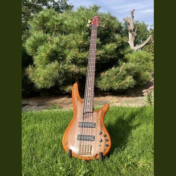 Dřevěná basová kytara s přírodním povrchem a hnědým hmatníkem. Na těle jsou dva černý humbuckery, čtyři ovládací knofíky a čtyři ladicí struny u most…