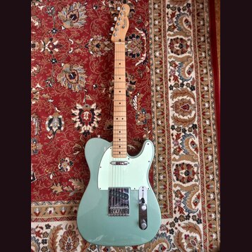 Světle zelená elektrická kytara typu Telecaster s javorovým hnedem. Na hlavě je viditelné logo Fender. Kytara má bílý pickguard a chromované kovové č…