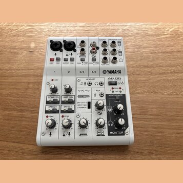 Bílá mixovací konzole značky YAMAHA, model AG06 MIXING CONSOLE. Zařízení obsahuje různé vstupy a výstupy, včetně konektorů XLR, jack 6,35 mm a USB. N…