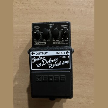Černý kytarový efekt pedál značky BOSS. Na horní straně jsou čtyři ovládací knoflíky s nápisy LEVEL, CHECK, VIBRATO a COSM. Na přední straně je bílá…