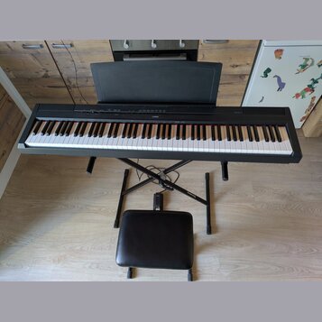 Černý digitální piano s klávesnicí, umístěné na černém X-stojanu s přiloženou černé polstrovanou židličkou. Na horní liště nástroje je uprostřed bílý…