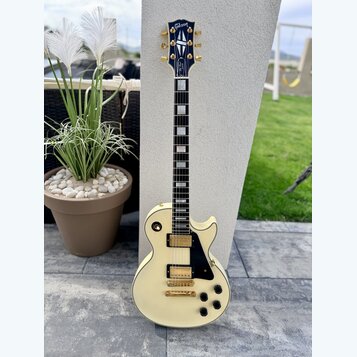 Elektrická kytara s krémovým povrchem korpusu a hmatníkem z tmavého dřeva s bílými blokovými vložkami. Na hlavě je zlatý nápis „Gibson“ a modrý ornam…