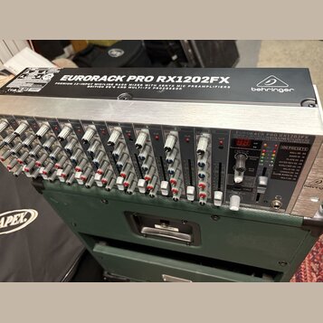 Stříbrný rackový mixážní pult s černým horním panelem. Na vrchu je bílý nápis EURORACK PRO RX1202FX a logo behringer. Přístroj obsahuje řadu knoflíků…