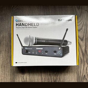 Fotografie znázorňuje krabici s bezdrátovým mikrofonním systémem. Na obalu je nápis CONCERT HANDHELD Frequency-Agile UHF Wireless System a logo SAMSO…