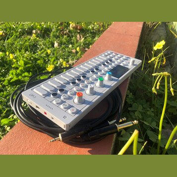 Bílý syntezátor OP-1 s klávesovou hrou a ovládacími knoflíky v různých barvách (modrý, zelený, oranžový). Na levé straně je viditelný nápis OP-1. Zař…