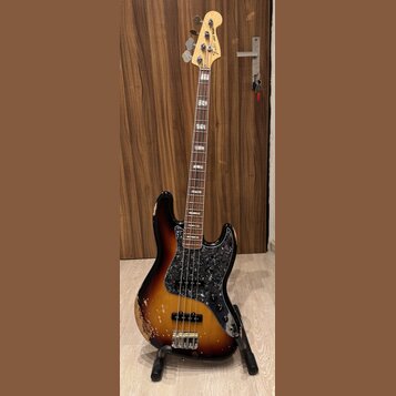 Elektrická basová kytara s trupem v barevnosti sunburst a černým pickguardem s texturou. Na hlavě hmatníku je nápis Fender Jazz Bass. Nástroj vykazuj…