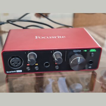 Červené audio rozhraní značky Focusrite. Na předním panelu je nápis Scarlett Solo a ovládací prvky s popisky 1, 2, 48V, AIR, MONITOR a DIRECT MONITOR…