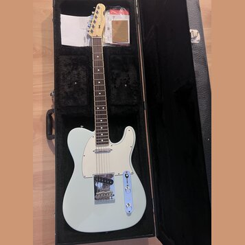 Světle modrá elektrická kytara typu Telecaster v černém tvrdém kufru. Na hlavě je viditelné logo Fender. Kytara má bílý pickguard, chromovaný most a…