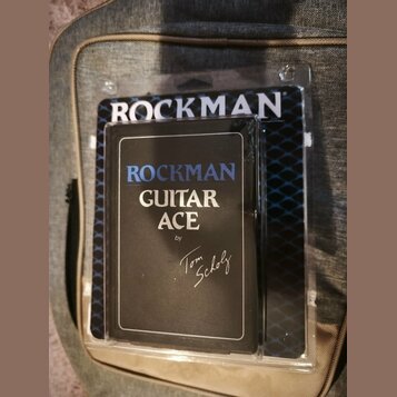 Kytarový procesor v černém obalu, který je umístěn v průhledném plastovém obalu. Na přední straně je bílý a modrý nápis ROCKMAN GUITAR ACE by Tom Sch…