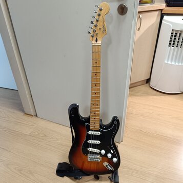 Elektrická kytara s trupem v barevnosti sunburst a černým pickguardem. Na hlavě hmatníku je viditelné logo Fender. Nástroj má světle hnědý krk a hmat…