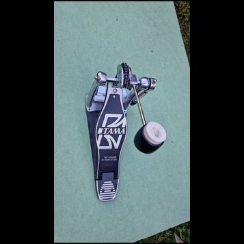 Černý basový pedál s kovovými mechanismy a bílým beaterem. Na stupni je bílé logo TAMA a nápis THE LEGEND IN INNOVATION. Povrch stupně vykazuje známk…