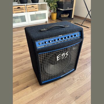 Černý basový kombo zesilovač s modrým ovládacím panelem a kovovou mřížkou. Na horní části je nápis EBS DROME PROFESSIONAL BASS COMBO 750 W a DEVELOPE…