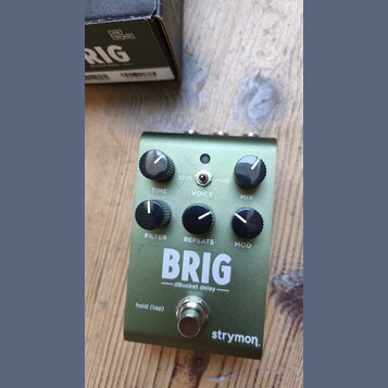 Zelený efekt pedál s nápisy BRIG a dBucket delay. Na spodní části je logo strymon. Pedál má šest černých otočných ovladačů s popisky TIME, VOICE, MIX…