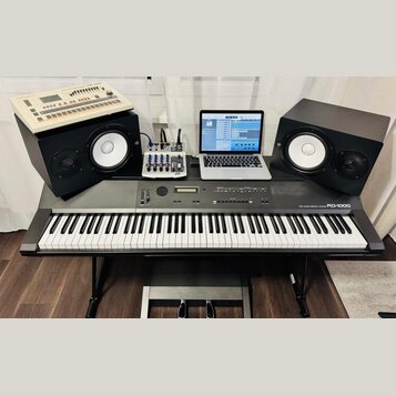 Digitální piano šedé barvy s nápisem ROLAND DIGITAL PIANO RD-1000, umístěné na černém stojanu s pedály. Na nástroji jsou umístěny dva černé studiové…