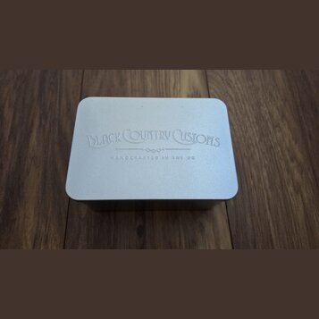Stříbrná kovová krabička s zaoblenými rohy. Na víčku je vyražen nápis BLACK COUNTRY CUSTOMS a pod ním menší text HANDCRAFTED IN THE UK. Povrch je mat…