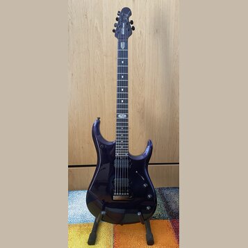 Tmavě fialová elektrická kytara s černým hmatníkem a dvěma humbuckery. Na hlavě je bílý nápis MUSIC MAN. Nástroj stojí na černém stojanu před dřevěno…