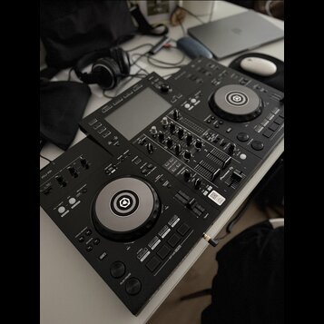Černý DJ 컨트롤러 s dvěma jogovými koly a centrálním mixážním sekcí. Na levé straně je viditelný nápis XDJ-RR a v pravé části logo Pioneer DJ. Za přístro…