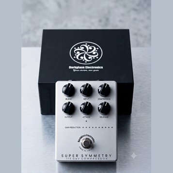 Stříbrný efektový pedál s šesti černými ovládacími knoflíky. Na spodní části je nápis SUPER SYMMETRY 115 GeV COMPRESSOR a kulaté logo Darkglass Elect…