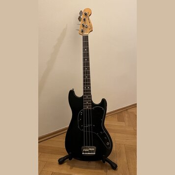 Černá elektrická basová kytara s hnědým hmatníkem a světlou dřevěnou hlavou. Na hlavě je nápis Fender MUSICMASTER BASS a logo Fender. Tělo má černý p…