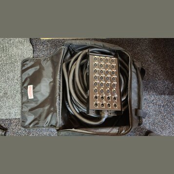 Černý rozpojitý kabel s konektory v kovovém boxu, umístěný v černé přepravní tašce. Na panelu jsou vidět početné konektory XLR a nápisy jako M1, M2,…