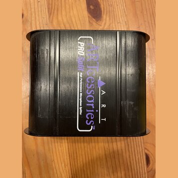 Černý obdélníkový box s žebrovaným povrchem. Na horní straně je bílý a fialový nápis ARTcessories TM, logo s trojúhelníkem a text A R T. Vedle něj je…