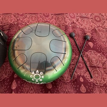 Kulatý kovový tongue drum s přechodem barvy od stříbrné v centru po zelenou na okrajích. Na spodní části nástroje je bílý nápis s písmeny A, G, E, C,…