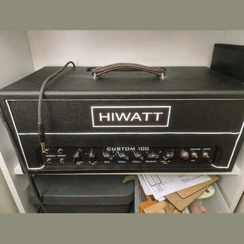 Černý kytarový zesilovač s koženou rukojetí a bílým lemováním. Na předním panelu je bílé logo HIWATT a nápis CUSTOM 100. Ovládací panel obsahuje přep…
