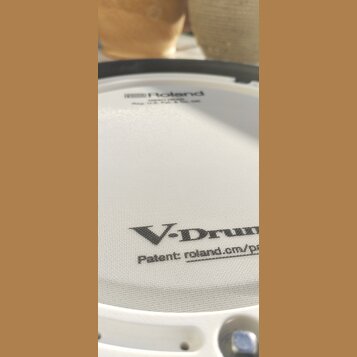 Bílá bubnová plocha s jemnou mřížkovou strukturou. V horní části je logo Roland a pod ním nečitelný text. Ve spodní části je velký nápis V-Drums a po…