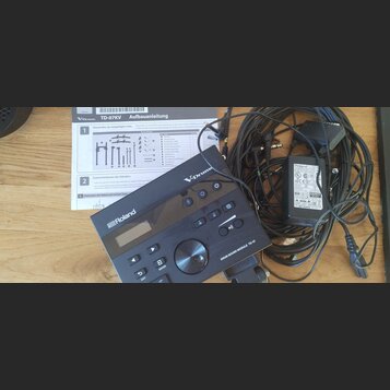 Na fotografii je černý elektronický bicí modul s nápisy Roland, V-Drums a DRUM SOUND MODULE TD-07. Vedle modulu jsou svázané černé kabely a černý nap…