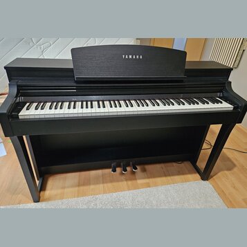 Černý digitální piano s klaviaturou a třemi pedály. Na horní části je nápis YAMAHA. Nástroj vykazuje známky běžného používání, jsou vidět drobné oděr…