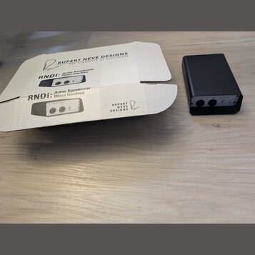 Na fotografii je černý, kompaktní box s dvěma vstupy a ovládacím prvkem. Vedle něj je otevřená kartonová krabice s nápisy RUPERT NEVE DESIGNS, The Si…