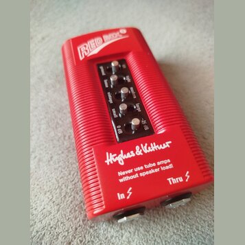 Červený DI box s rýhovaným povrchem. Na horní části je nápis RED BOX a v dolní části logo Hughes & Kettner s varovným textem Never use tube amps with…