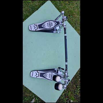 Dvojpedál na bicí soupravu v černé a chromované barvě. Na obou pedálech je bílý nápis IRON COBRA 200 a logo TAMA. Nástroj vykazuje známky běžného pou…