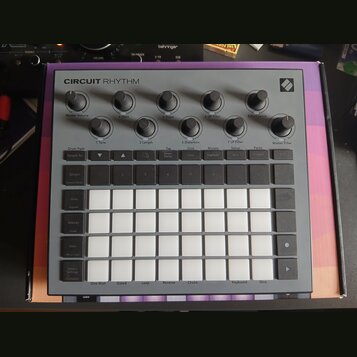 Šedý sampler Novation Circuit Rhythm s bílými podsvícenými pady v mřížce 4x8. V horní části jsou umístěny otočné ovladače s popisky Master Volume, Tu…