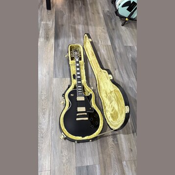 Černá elektrická kytara s tvarem single-cutaway, umístěná v otevřeném černém kufru s žlutým vnitřkem. Na hlavě kytary je viditelné logo značky Gibson…