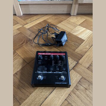 Černý efekt pedál s červeným pruhem a nápisy TC-HELICON harmony GXT a VOICETONE™. Zařízení obsahuje několik ovládacích knoflíků, přepínačů a displej…