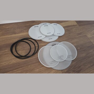 Sada bílých bubnových blan v různých velikostech a tři černé gumové prstence. Pět blan nese nápisy drum-tec a real feel, další čtyři mají nápis Mille…