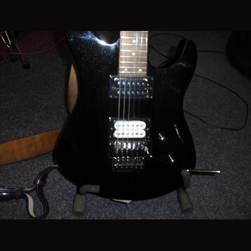 Černá elektrická kytara s třemi snímači (dvě humbuckery a jeden single-coil). Kytara má hnědý dřevěný hриф a kovový tremolo systém s páčkou. Na těle…