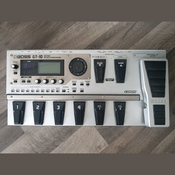 Stříbrný kytarový efektový procesor s nápisem BOSS GT-10 GUITAR EFFECTS PROCESSOR. Zařízení má digitální displej, několik ovládacích tlačítek a v pra…